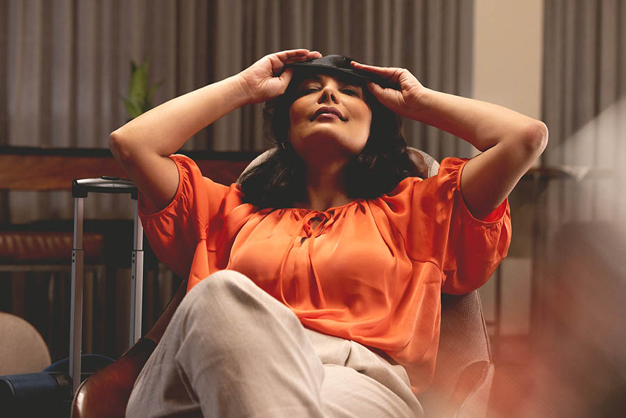 A woman sitting in a chair with her hands on her head, relaxing with an eye mask, before her flight.