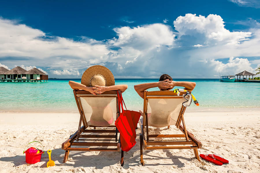 image of couple from behind sat in deck chairs on a white sand beach looking out to turquoise ocean.